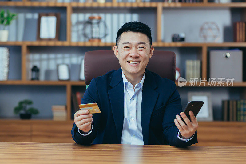 Happy Asian working in the office, holding a bank credit card and phone for online shopping, looking at the camera and smiling